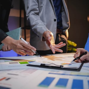 Personas reunidas en una mesa revisando gráficos y documentos con datos financieros y de análisis, señalando información con bolígrafos en un entorno profesional.
