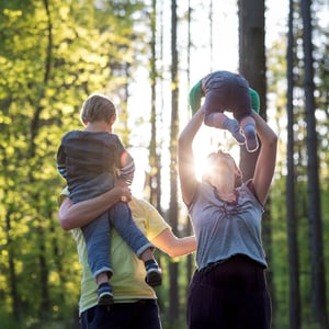 Dos personas adultas sosteniendo a dos niños pequeños, son una familia, en un bosque iluminado por luz natural, con árboles altos y follaje verde en el fondo.