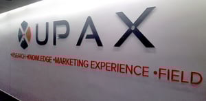 Logotipo de UPAX en una pared blanca, acompañado de las palabras Research, Knowledge, Marketing Experience y Field en letras rojas.