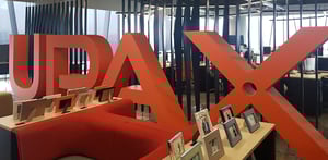 Área de oficina moderna con un gran letrero rojo que forma la palabra UPAX, acompañado de muebles y marcos decorativos sobre una mesa, con estaciones de trabajo al fondo.