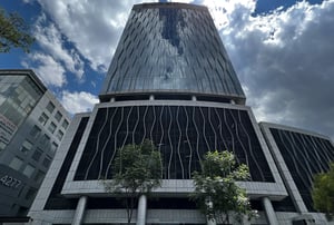Edificio del Grupo UPAX moderno con fachada de vidrio y diseño ondulado en los niveles inferiores, rodeado de árboles y otras construcciones, bajo un cielo parcialmente nublado.