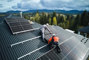 Instalación de paneles solares en el techo de una estructura metálica, con dos técnicos trabajando en el montaje, rodeados de un paisaje montañoso y bosques.