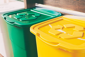 Contenedores de reciclaje en colores amarillo, verde y blanco, alineados bajo una estructura de madera, cada uno con el símbolo de reciclaje en la tapa.