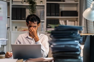 Persona frente a una computadora en oficina, rodeada de carpetas y pilas de documentos.