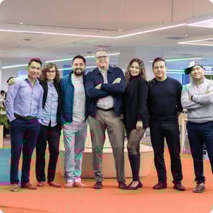 Grupo de personas posando juntas en un espacio de oficina moderno con alfombra naranja, mobiliario minimalista y luz blanca, transmitiendo trabajo en equipo y colaboración.