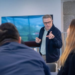 Persona de pie frente a una pantalla grande en una sala de reuniones, gesticulando mientras presenta información a un grupo sentado, en un entorno corporativo moderno.
