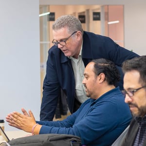 Personas colaborando en una oficina moderna, una de ellas gesticulando mientras conversa con otra que se inclina sobre el escritorio, con fondo de paredes claras y mobiliario contemporáneo.