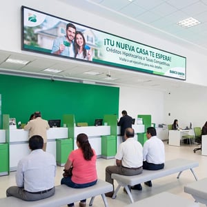 Sucursal bancaria con clientes en ventanillas y pantalla superior que promociona créditos hipotecarios.